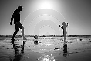 Father and son play soccer or football on the beach on summer family holidays. Dad and child playing outdoor, silhouette