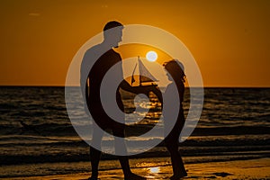 Father and son playing with toy sailboat, toy ship yacht on sea beach at summer sunrise. Sunset silhouette of father and