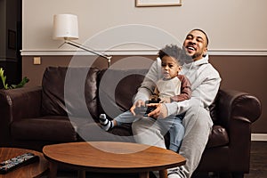 Father and son playing a video game together at home. Young man and little boy sitting on sofa holding joysticks and having fun