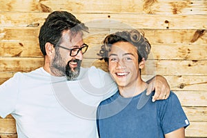 Father and son portrait - adult and young teenager generations smile and hug together with wooden background - youthful and mature