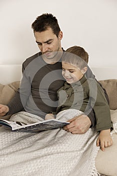 Father and son reading a book on the bed at home. Young attractive man and little boy resting in bedroom. Natural earth