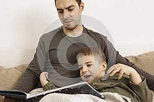 Father and son reading a book on the bed at home. Young attractive man and little boy resting in bedroom. Natural earth
