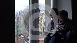 Father and son are reading a book and compose a story from pictures in it. Wall and the window are in background