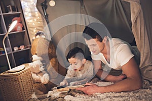 Father and son are reading book with magnifying glass at night at home.