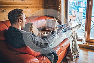 Father and son reading book together lying on the cozy sofa in warm country house. Reading to kids conceptual image