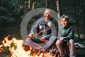 Father and son roast marshmallow candies on the campfire in fore