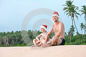 Father and son in santa hats sit together on sandy tropical beach