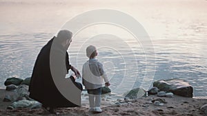 Father and son on the shore, little boy throw stone into the water. Man squatting and show boy how play stone skipping.