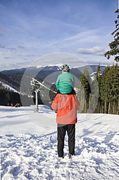 Father with son on shoulders stands against the backdrop of mountains, cable lift and coniferous forest. Winter day. Blue sky