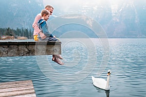 Father with son sit on wooden pier and look on white swan swims