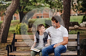 Father with son sitting on bench outdoors in town, reading a book.