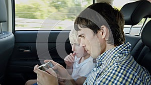 Father with son sitting in car seat and playing on smart phone while having car ride in backseat