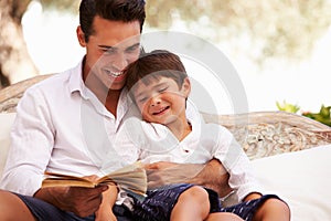 Father And Son Sitting In Garden Reading Book Together