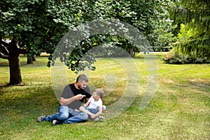 Father and son are sitting on the grass in park outdoor. Father teaches son, tells a story. Kindred love. Affection. Trusting