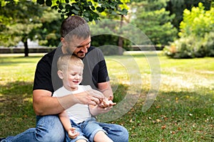 Father and son are sitting on the grass in park outdoor. Father teaches son, tells a story, plaing. Kindred love. Affection.