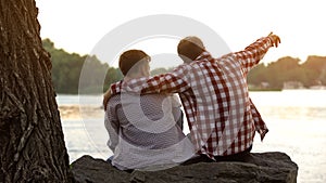 Father and son sitting on river bank, dad pointing at distance, enjoying view