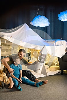 Father and son sitting together in blanket fort and playing with toy rocket