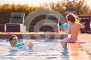 Father And Son Squirting Each Other With Water Pistols Playing In Swimming Pool On Summer Vacation