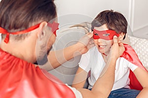 Father and son in superheroe costumes at home sitting on sofa man putting mask on boy smiling close-up photo