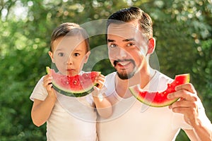 Father and son together eating watermelons, both man and kid are holding slices of juicy watermelons, green leaves in background photo