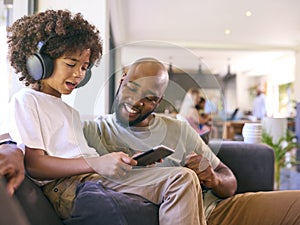 Father And Son Using Digital Tablet With Headphones At Home With Multi-Generation Family Behind