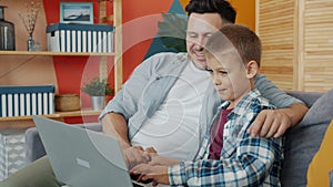 Father and son using laptop computer talking sitting on couch at home together