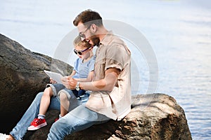 father and son using tablet near lake