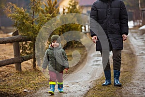 Father and son walking in the fresh air in rubber boots on the puddles and mud after the rain. Little child holding hand
