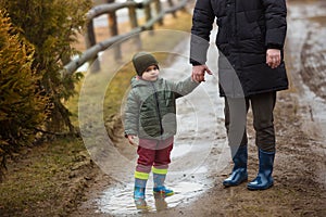 Father and son walking in the fresh air in rubber boots on the puddles and mud after the rain. Little child holding hand