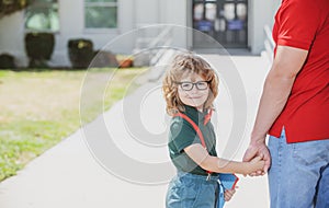 Father and son walking trough school park. Portrait of happy nerd pupil holding teachers hand.