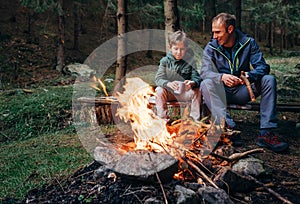 Father with son warm near campfire, drink tea and have conversation