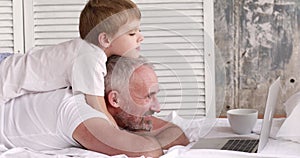 Father and son watch cartoons on a laptop lying on the bed.