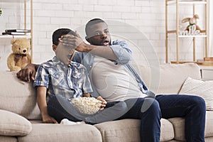 Father and son watching scary movie together, dad covering kid`s eyes