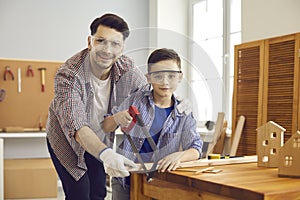 Father and son wearing safety goggles while sawing wood plank at carpenter's workshop