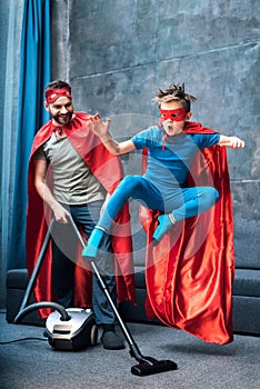 Father in superhero costume vacuuming carpet while son in superhero costume jumping