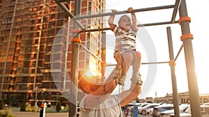 Father supporting little son while she going across monkey bars on outdoor workout playground on sunny day.