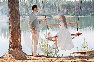 A father swings his daughter on a swing on the shore of a forest lake