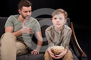 Father talking and gesturing to little son sitting on sofa with popcorn in bowl