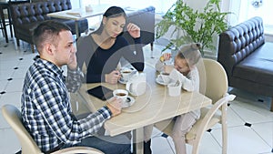 Father talking to phone while sitting with his wife and daughter in cafe or restaurant. Mother looking angry at him.