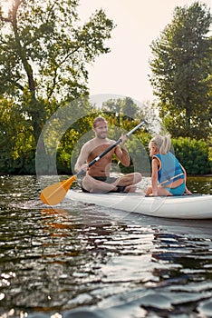 Father teaching his child, little daughter sup surfing on a river surrounded by the beautiful nature on a summer day