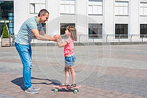 A father is teaching his daughter skateboarding in a city park. A girl is learning balancing on a board. Happy family