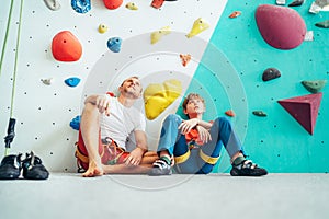 Father and teenage son sitting near the indoor climbing wall. They resting after the active climbing. Happy parenting concept