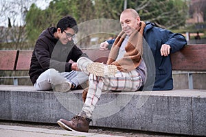 Father and teenage son talking and laughing sitting on a bench in a park.
