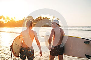 Father with teenager son walking with surfboards by sandy ocean beach with palm trees on background lightened with sunset sun They