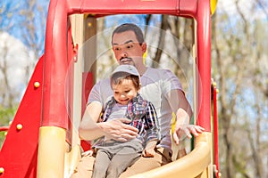 Father and toddler son sliding from children slide in the park. Child is sitting on knees of dad, father is funny and scared, kid