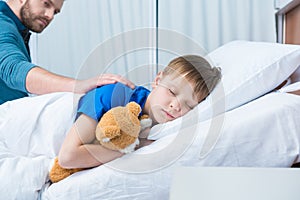 Father touching sick little son lying in hospital bed with teddy bear