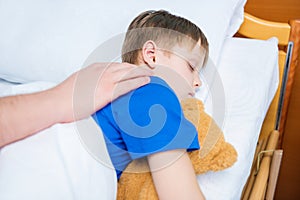 Father touching sick little son lying in hospital bed with teddy bear