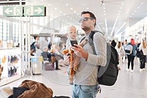 Father traveling with child, holding his infant baby boy at airport terminal, checking flight schedule, waiting to board