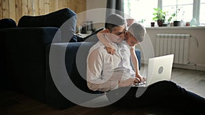 Father is trying to work on laptop with his son cuddling him in living room.