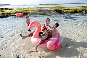 Father and two daughters ride flamingo buoy on the beach